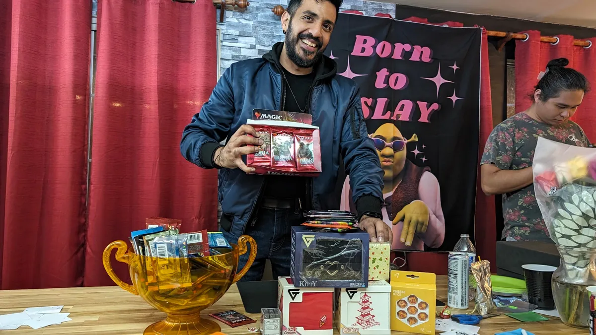 Guild host showing Magic booster packs at a prize table with trophies and collector items